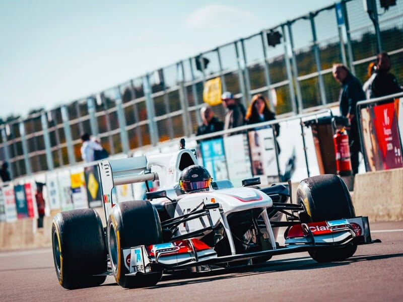 Pilote en Formule 1 blanche dans la voie des stands du circuit de Magny-Cours avec des spectateurs en arrière-plan.