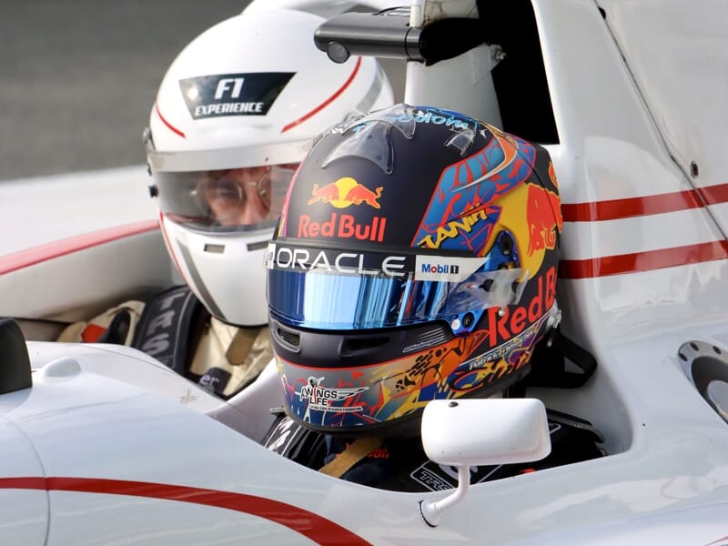 Pilote portant un casque Red Bull et un instructeur en casque blanc dans une monoplace de Formule 1 blanche.