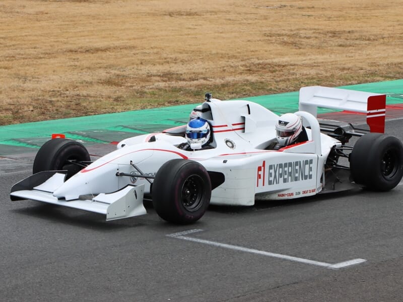 Monoplace blanche de Formule 1 avec deux personnes à bord sur un circuit, marquage F1 Experience visible.