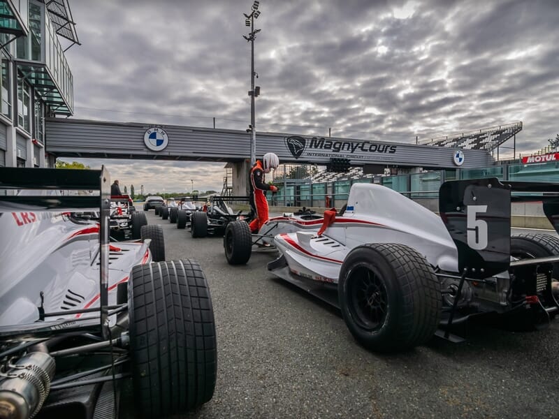 Voitures de course de Formule Renault alignées sur la piste du circuit de Magny-Cours sous un ciel nuageux.