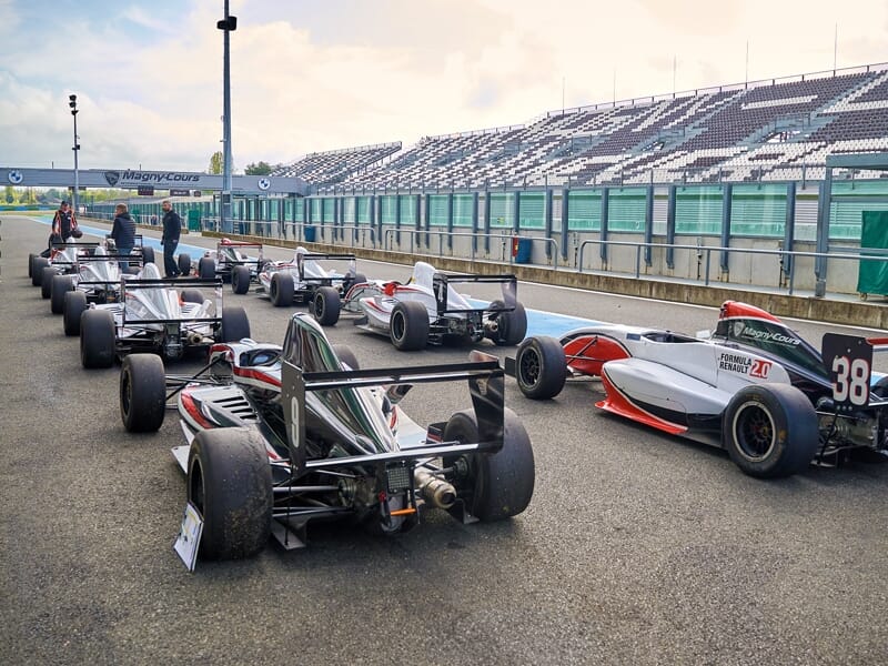 Plusieurs voitures de Formule Renault stationnées sur la piste du circuit de Magny-Cours avec des spectateurs en arrière-plan.