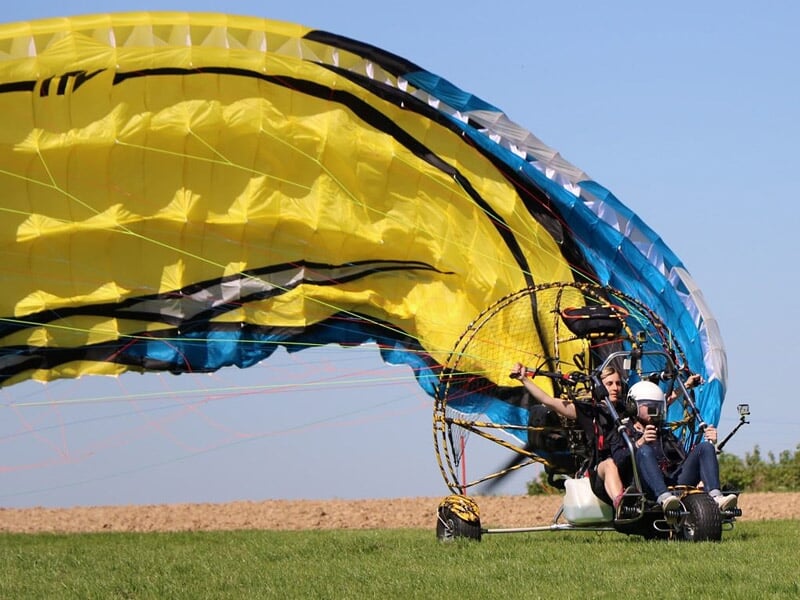 Paramoteur au sol avec voile jaune et bleue déployée prêt à décoller sur un terrain herbeux sous un ciel clair.