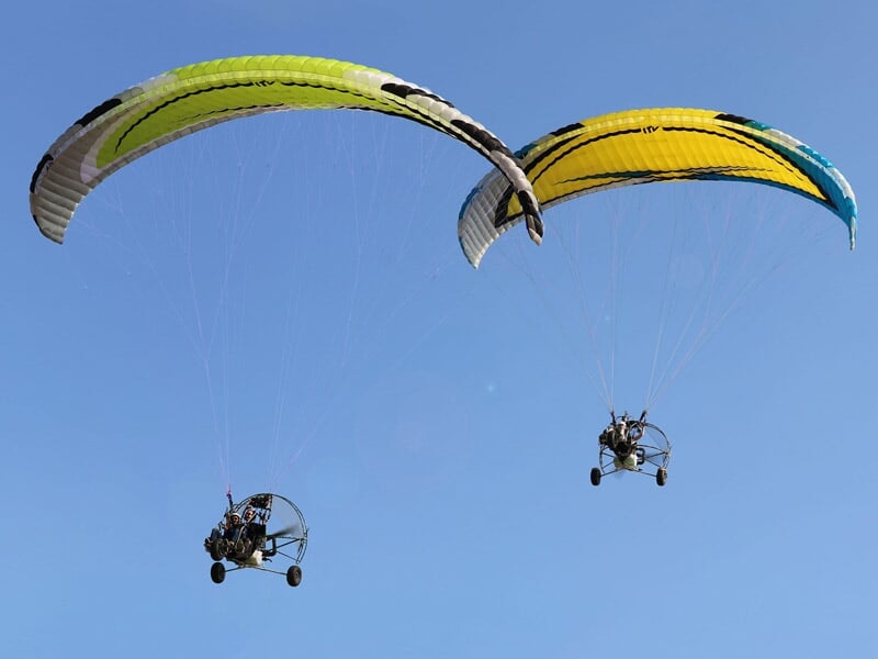 Deux paramoteurs en vol avec voiles jaune et verte, évoluant côte à côte dans un ciel bleu dégagé.