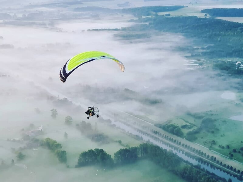 Paramoteur volant au-dessus d'un paysage brumeux avec arbres et champs visibles à travers la brume matinale.