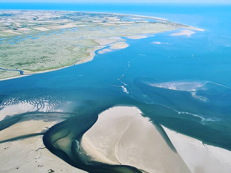 Vue aérienne d'un estuaire avec plages de sable, eaux calmes et zones humides sous un ciel clair.