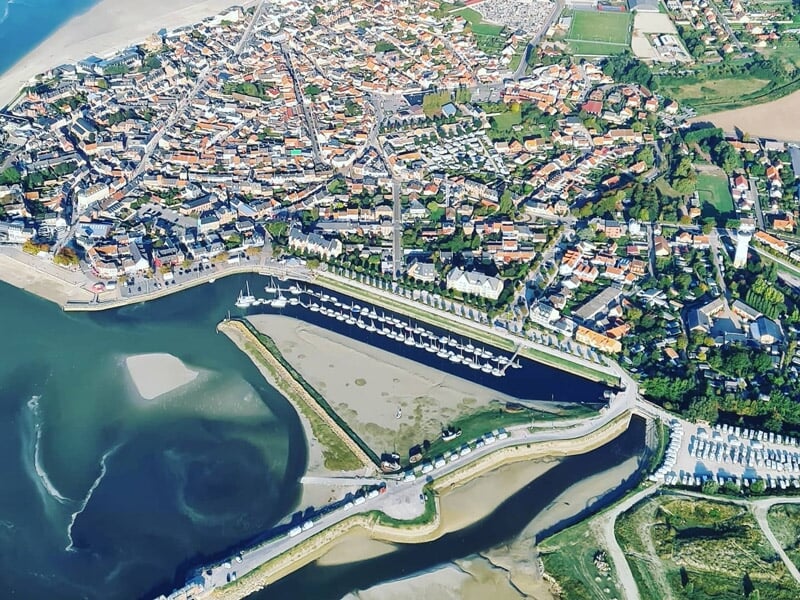 Vue aérienne d'une ville côtière avec un port de plaisance, des bâtiments et des espaces verts environnants.