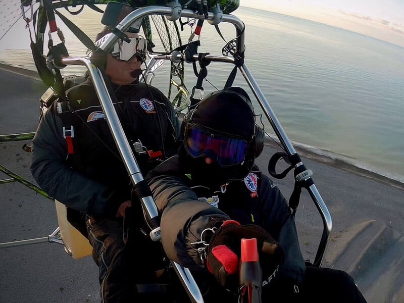 Deux personnes en paramoteur, équipées de casques et lunettes, prêtes à décoller ou en vol au-dessus de la mer.