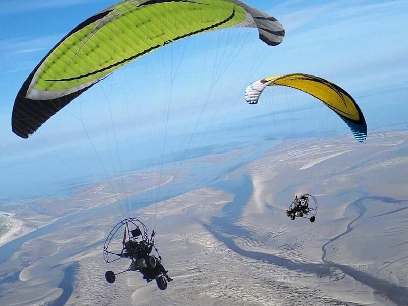 Deux paramoteurs volant au-dessus d'un paysage côtier avec plages, dunes et eaux peu profondes visibles en contrebas.