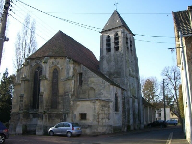 Église médiévale en pierre avec un clocher et des vitraux, située dans un village, entourée de voitures garées, sous un ciel bleu clair.