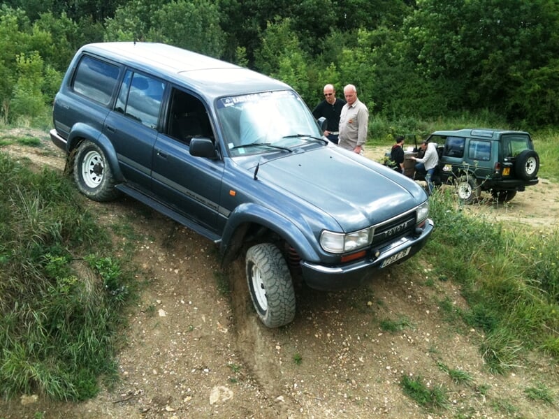 4x4 gris sur une pente boueuse, deux hommes discutant à proximité, d'autres véhicules et personnes en arrière-plan, lors d'une sortie en tout-terrain.