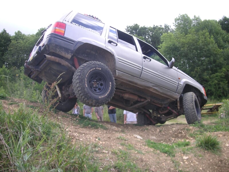 4x4 gris foncé en position inclinée sur une colline boueuse, ses roues arrière soulevées du sol, démontrant ses capacités de franchissement tout-terrain.
