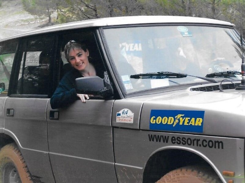 Femme souriante à bord d'un 4x4 gris avec des autocollants sponsorisés, conduite hors-piste sur un chemin boueux, entourée de végétation.