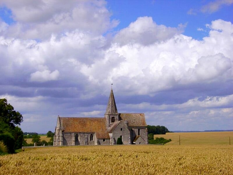 Église rurale en pierre avec un clocher et un toit en tuiles, entourée de champs de blé sous un ciel partiellement nuageux, créant une scène pittoresque.