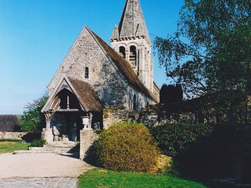 Église médiévale en pierre avec un clocher pointu et un porche en bois, située dans un jardin avec des buissons et des arbres, sous un ciel bleu clair.