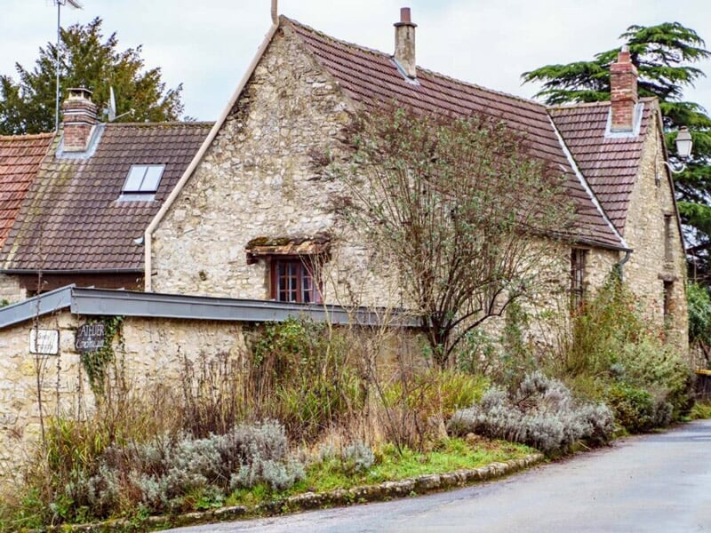 Vieille maison en pierre avec un toit en tuiles et des volets en bois, entourée de végétation sauvage, située au bord d'une route de campagne.