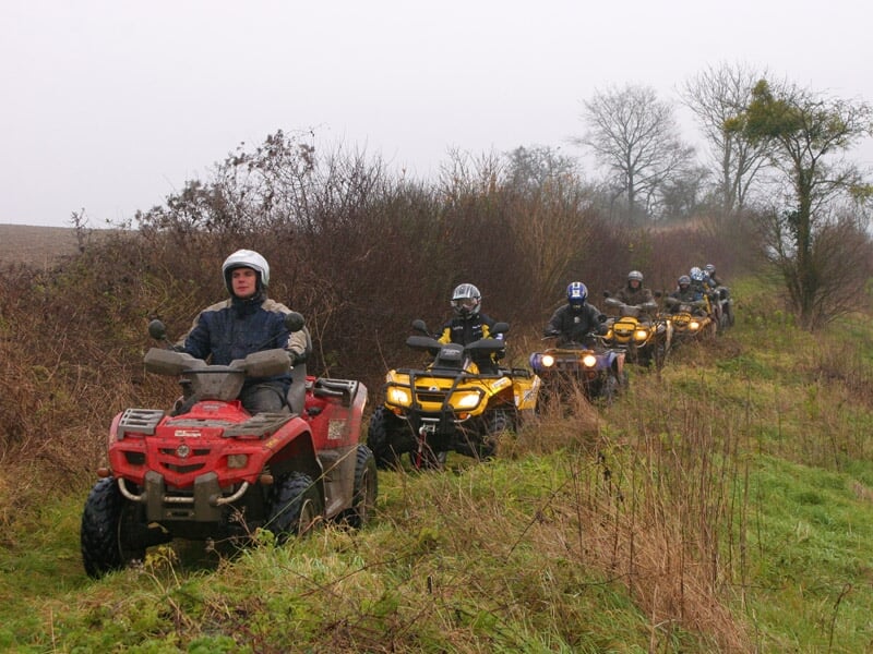 File de quads avançant sur un sentier en pleine nature par temps gris, les conducteurs portant des casques et des vêtements de protection. Le quad rouge en tête est suivi de plusieurs quads jaunes.