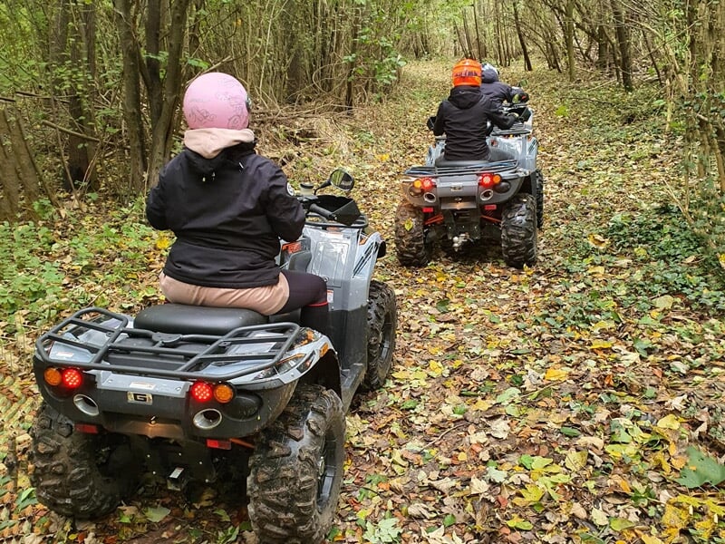 Deux personnes en quad traversant un sentier forestier couvert de feuilles mortes, portant des casques rose et orange. Les quads noirs contrastent avec la verdure environnante.