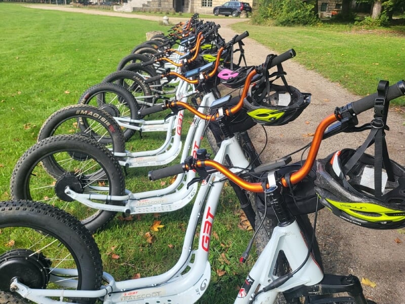 Ligne de trottinettes électriques tout-terrain alignés sur une pelouse verte, avec des casques accrochés aux guidons. Les vélos blancs avec des accents orange sont prêts pour une activité de plein air