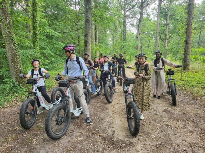 Groupe en forêt, équipés de trottinettes électriques tout-terrain, souriants et posant ensemble parmi les arbres. Les casques et les vêtements de sport suggèrent une randonnée en plein air.