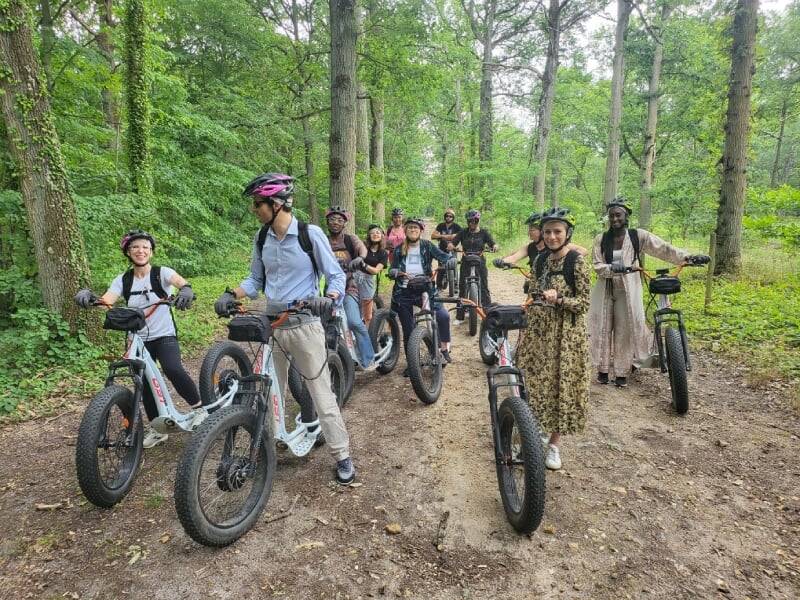 Groupe en forêt, équipés de trottinettes électriques tout-terrain, souriants et posant ensemble parmi les arbres. Les casques et les vêtements de sport suggèrent une randonnée en plein air.