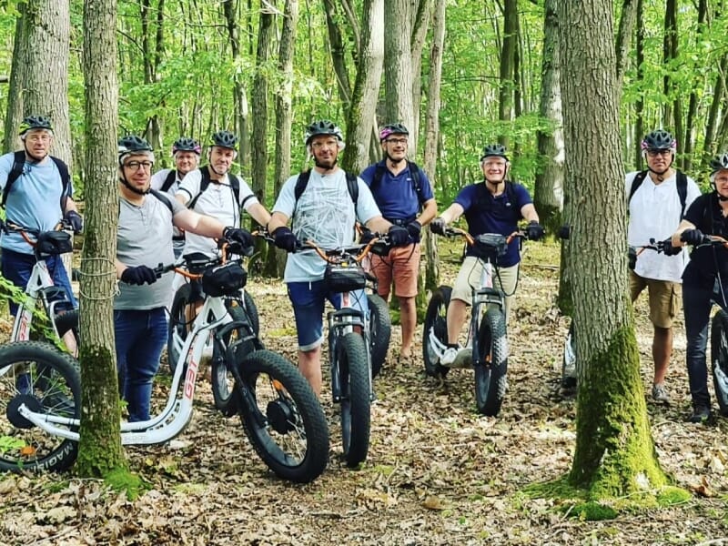 Groupe de personnes en trottinette électrique tout-terrain dans une forêt, portant des casques et des sacs à dos, prêts pour une balade en nature. Les arbres verts et le sentier terreux forment un cad