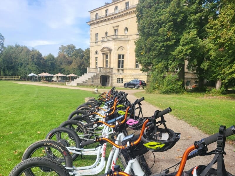 Alignement de vélos tout-terrain avec casques colorés devant un manoir historique en pierre, entouré de verdure et d'arbres. Journée ensoleillée avec ciel partiellement nuageux, tables et chaises sous