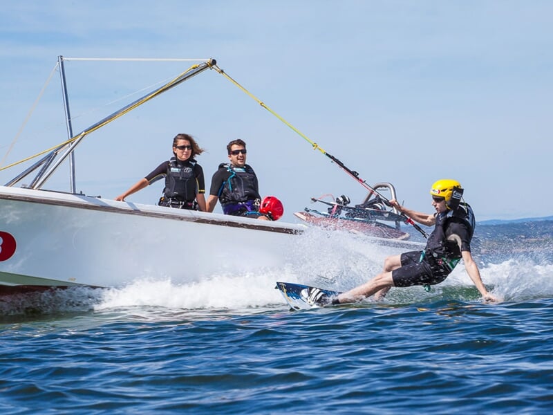 Personne en combinaison et casque jaune fait du kitesurf tracté par un bateau sur l'eau bleue du Bassin de Thau.