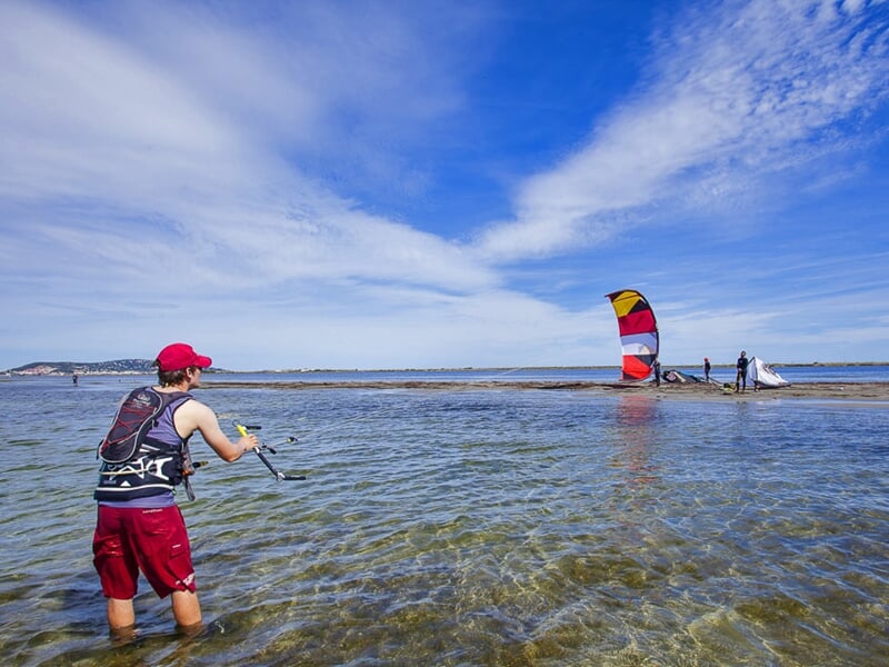 Jeune homme en short rouge et gilet de sauvetage pratique le kitesurf dans une eau peu profonde sous un ciel bleu.