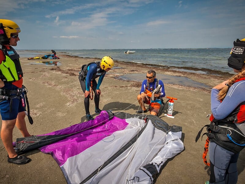 Groupe de personnes en combinaison prépare une voile de kitesurf violette et grise sur une plage sableuse.