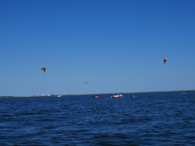 Plusieurs kitesurfeurs évoluent sur le Bassin de Thau sous un ciel bleu sans nuages.