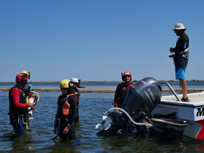 Quatre personnes en combinaison et casque jaune discutent dans l'eau près d'un bateau à moteur.