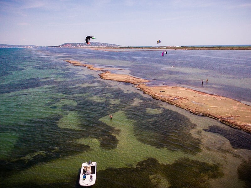 Vue aérienne du Bassin de Thau avec un bateau blanc et plusieurs kitesurfeurs sur l'eau turquoise.