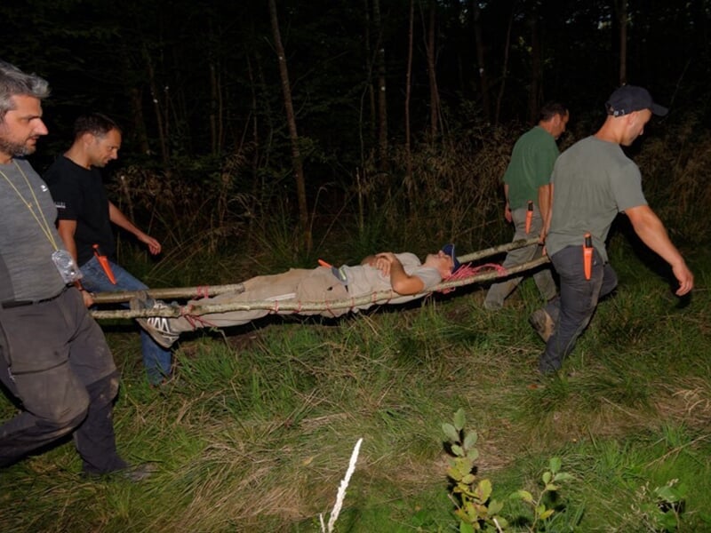 Quatre hommes transportant une civière avec une personne allongée, en pleine forêt, lors d'un exercice.