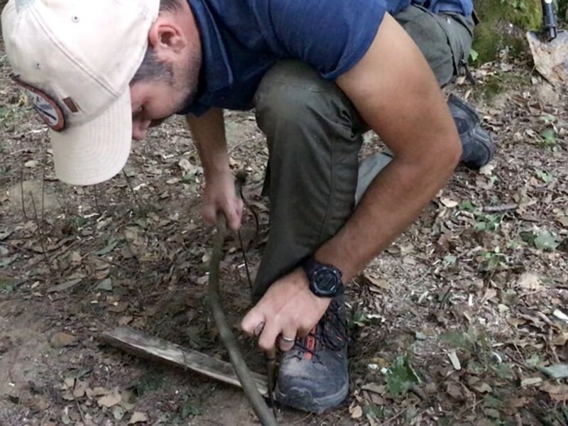 Homme en pleine forêt utilisant un arc à feu pour allumer un feu, vêtu de vêtements de survie avec une montre robuste, entouré de feuilles et de bois au sol