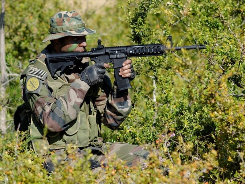 Soldat en tenue camouflage, armé, en position de tir dans un environnement boisé et dense.