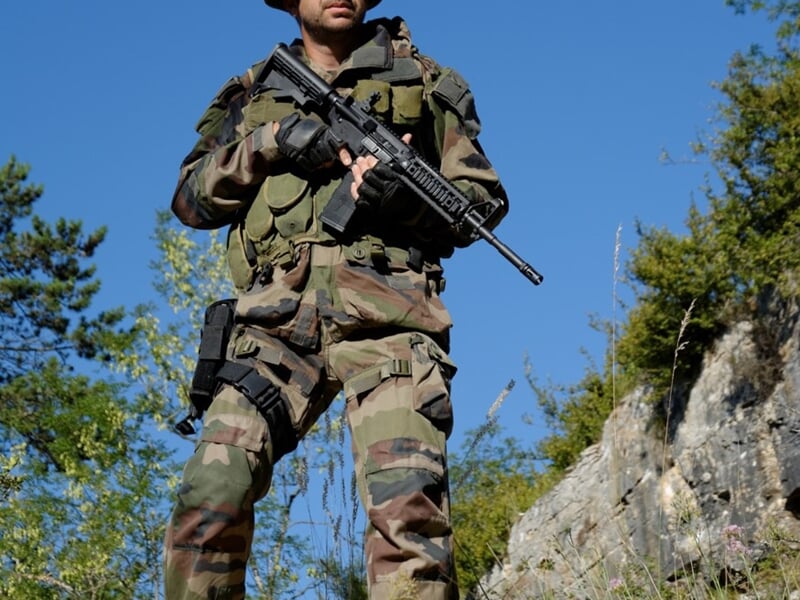Militaire en uniforme complet avec arme, debout sur un chemin rocheux sous un ciel bleu clair.