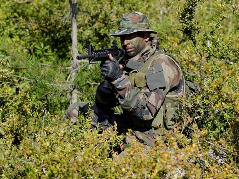 Militaire en tenue de camouflage, armé, accroupi dans un buisson dense lors d'un exercice.