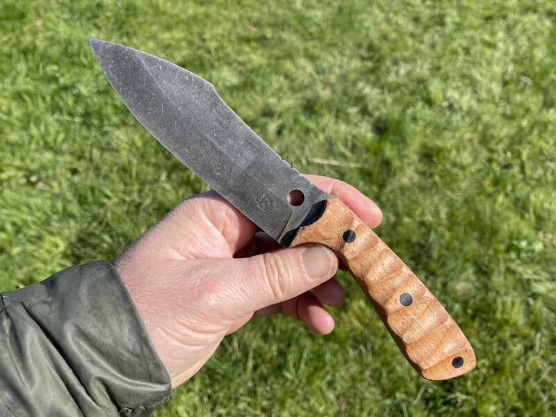 Couteau de survie avec lame en acier brut et manche en bois texturé, tenu par une main gauche vêtue d'une veste kaki, fond d'herbe verte en extérieur