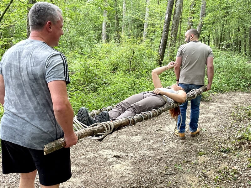 Deux hommes transportent une femme sur une civière improvisée avec des branches, en pleine forêt, lors d'une formation de secourisme en pleine nature