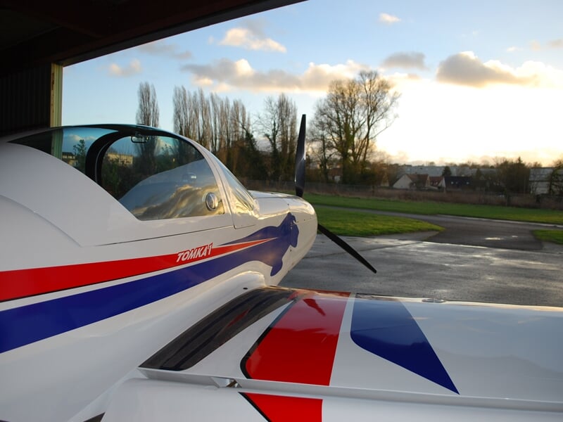 Vue rapprochée de l'aile et du cockpit d'un avion léger blanc avec des bandes rouges et bleues au sol.
