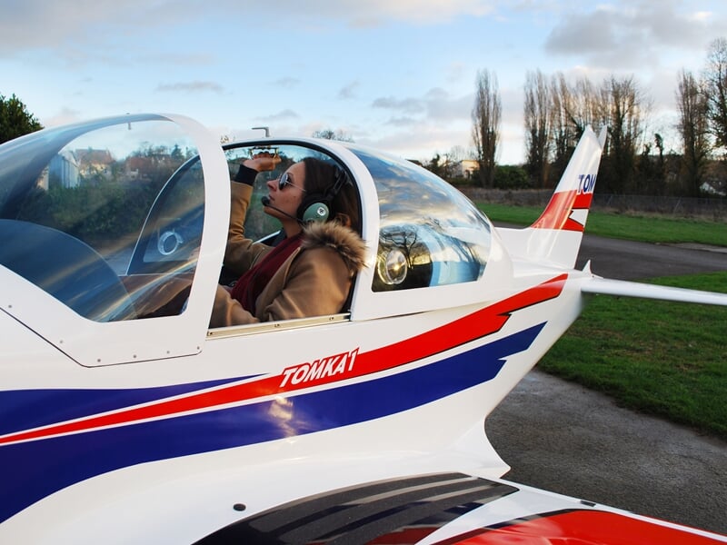 Personne assise dans le cockpit d'un avion léger blanc avec casque et veste, prêt au décollage.