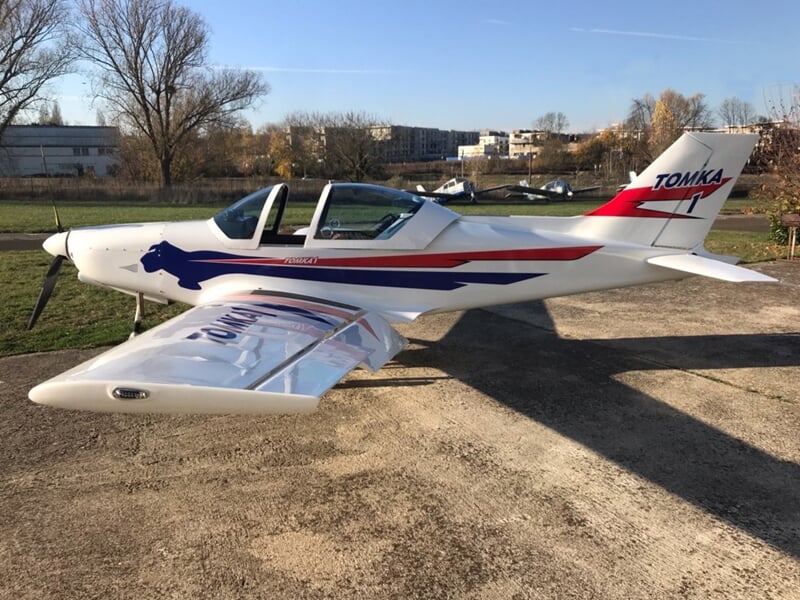 Avion léger blanc avec décorations rouges et bleues stationné sur une surface en gravier, arbres et bâtiments visibles.