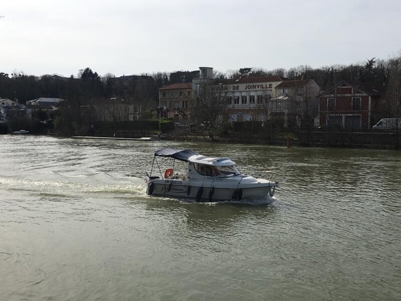 Bateau à moteur blanc avec canopy bleu naviguant sur une rivière devant le bâtiment « Mairie de Joinville », rive boisée en arrière-plan.