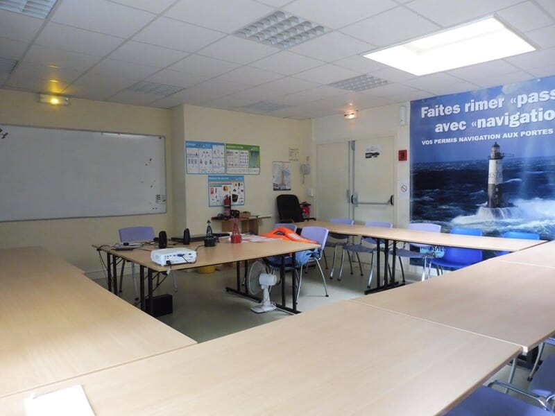 Salle de formation nautique avec tables en U, chaises violettes, tableau blanc, affiches pédagogiques et grande affiche murale sur la navigation.