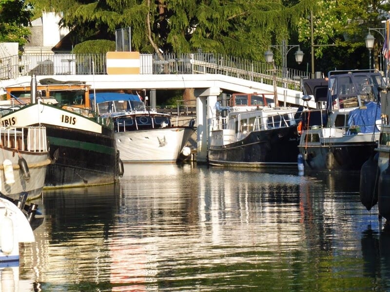 Port fluvial bordé de péniches et bateaux de plaisance amarrés, pont piéton blanc en arrière-plan, reflets sur l’eau calme.