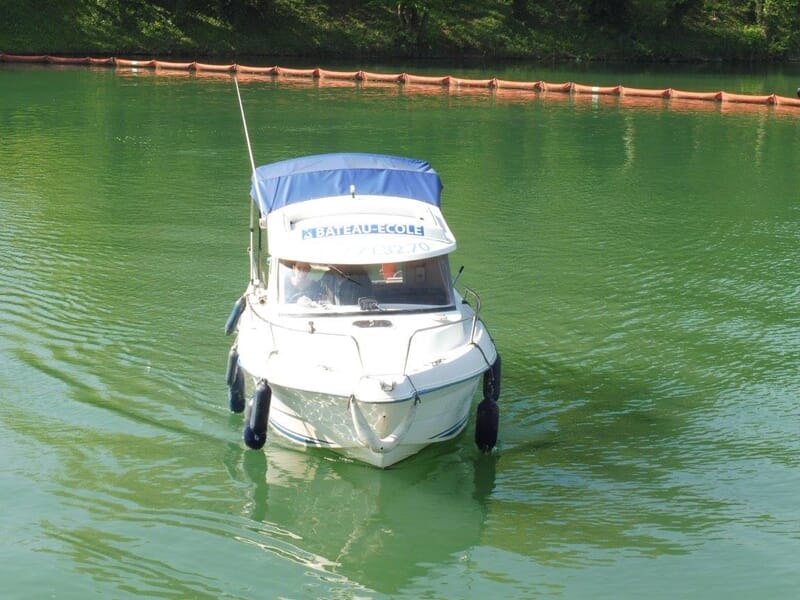 Bateau-école blanc à canopy bleu avançant sur une eau verte, panneau « BATEAU ECOLE » visible sur le toit, pilote à bord.