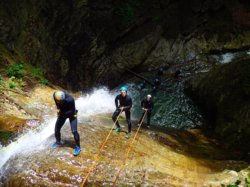 Groupe de personnes équipées de casques et combinaisons noires descendent en rappel sur une cascade en canyoning.
