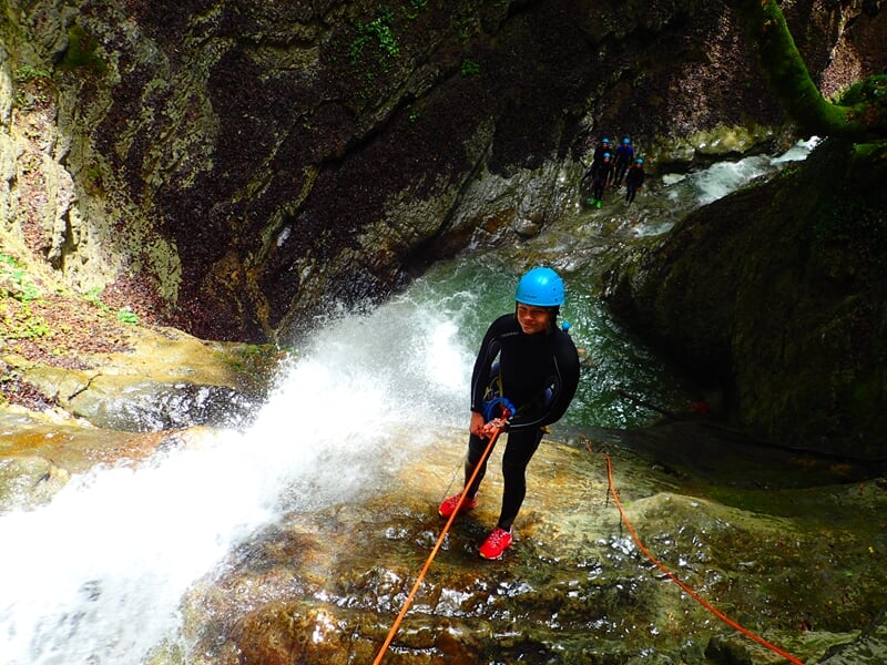 Personne en combinaison noire et casque bleu descend en rappel le long d'une cascade dans un canyon verdoyant.