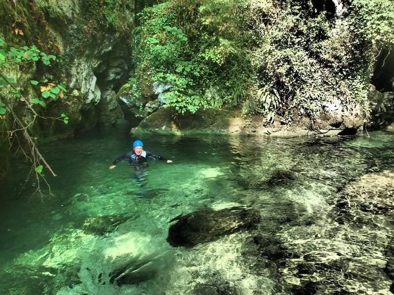 Personne en casque bleu nage dans une eau claire et calme entourée de rochers et de végétation dense.