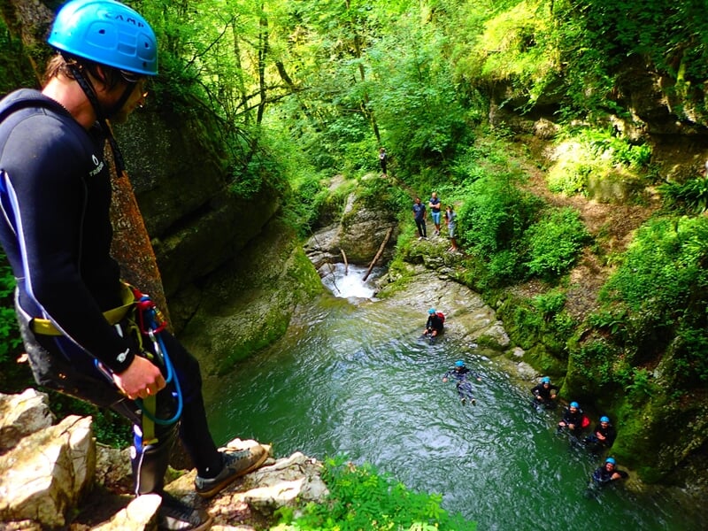 Personne en combinaison noire observe un groupe de personnes dans un canyon avec une eau verte et des rochers.